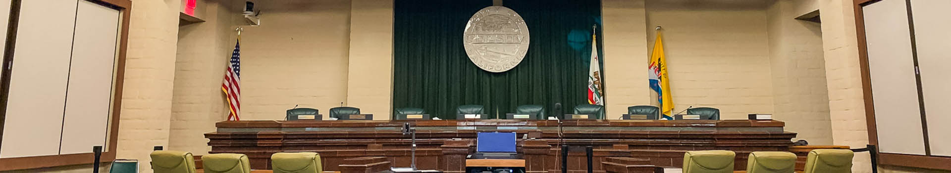 Clerk Home Banner of empty council chambers with light and dark green leather chairs, Folsom emblem hanging, American Flag, California Flag, and Folsom Flag