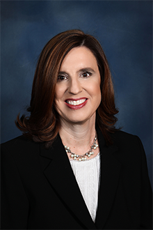 Elaine Andersen professional portrait headshot with a navy blue background and wearing a black blazer with a white top and a necklace