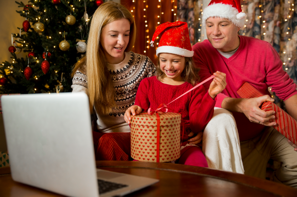 Getty Image of a mother, father, and child opening a present in winter clothes, and the dad and daughter wearing red and white santa hats, with a silver laptop in front of them and christmas decorations in the background