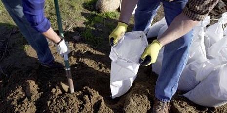 Photo of 2 people wearing gloves, filling sandbags with the person on the left shoveling sand while the person on the right holds the sandbag open for filling