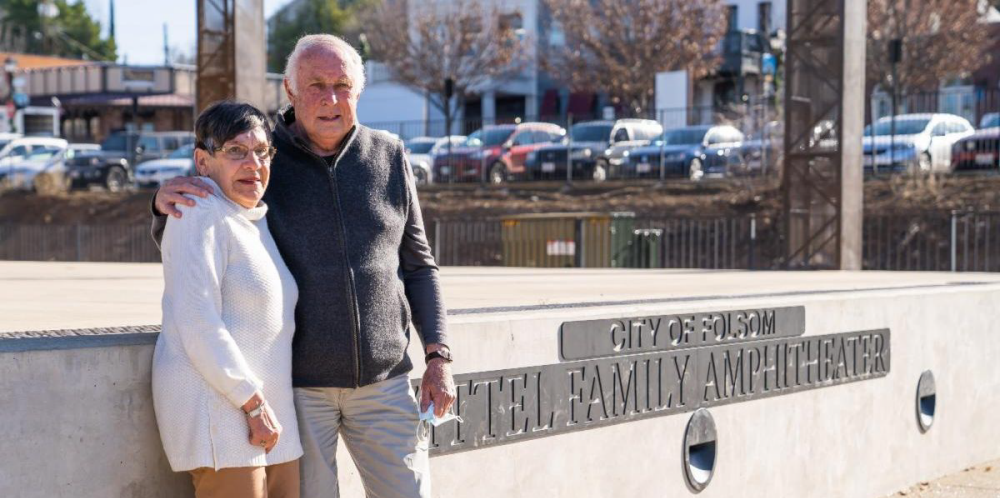 Photo of Zittel family in front of the City of Folsom Zittel Family Amphitheater with Historic District businesses and cars in the background. The woman on the left is wearing a white sweater with tan pants and glasses, and the man on the right is wearing a gray zip-up sweater with light gray pants.