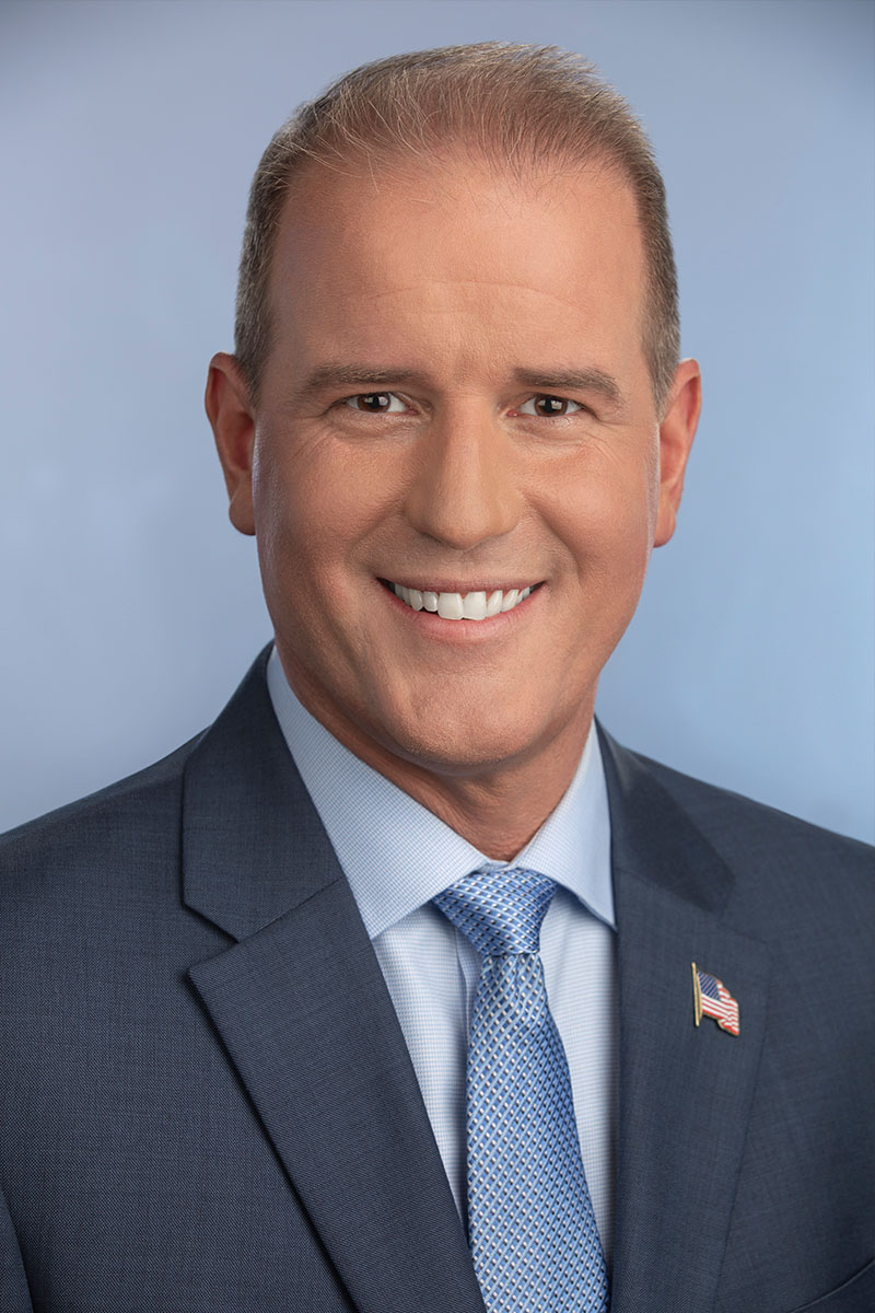 Councilmember Mike Kozlowski wearing a dark navy suit with a light gray button up shirt, a blue tie, and an American Flag pin formal portrait headshot with a light blue background