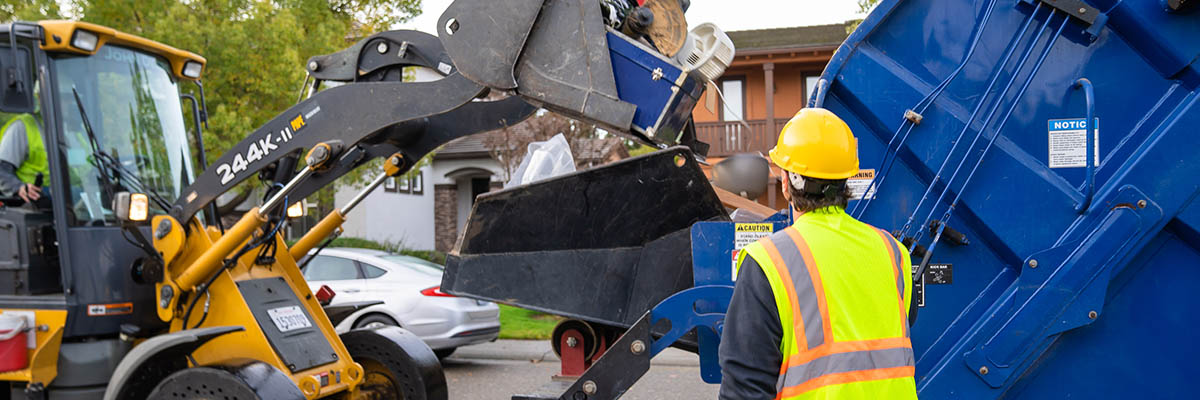 Waste Banner, a bulldozer placing large items into the back of a blue refuse truck with an employee wearing a reflective vest and hardhat watching