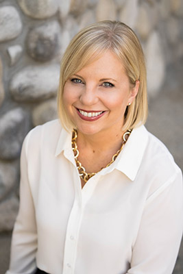 Communications Director Christine Brainerd in a casual headshot wearing a white button-up blouse and a gold chain necklace with gray cobblestone in the background