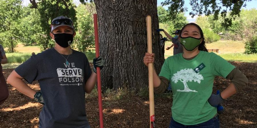 photo of two people holding rakes and wearing masks with other volunteers, a large tree, and open grassy areas in the background. Volunteers are wearing a blue CSD shirt and a green tree shirt