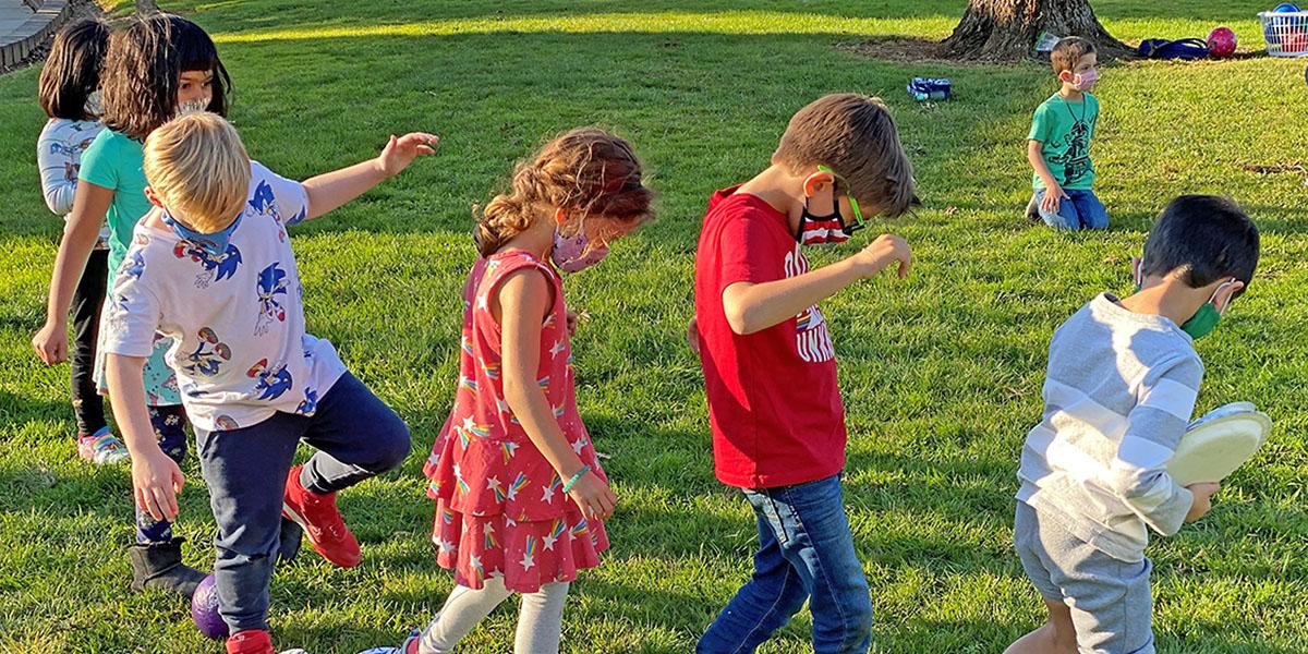 Summer Day Camp photo of 6 children walking in a line while wearing masks, and another child kneeling on the grass in the background