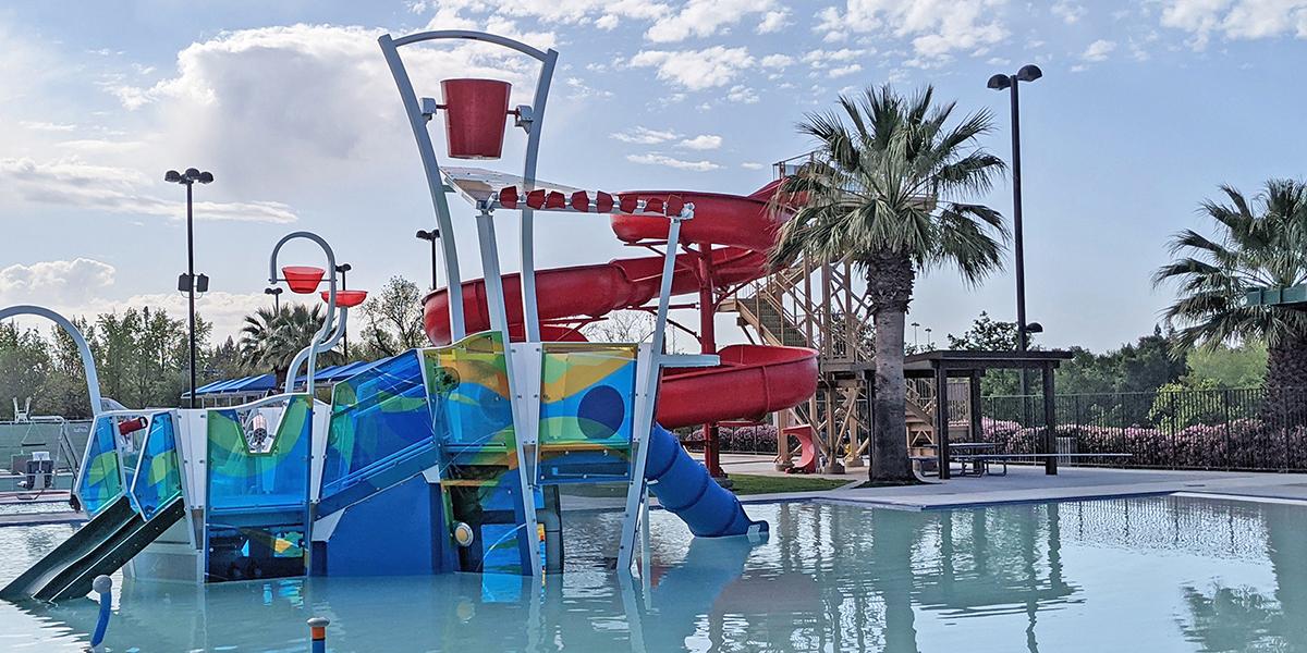 Aquatic Play Structure with a kid pool surrounding it and a red slide behind it. There are trees, blue sky, and clouds in the background