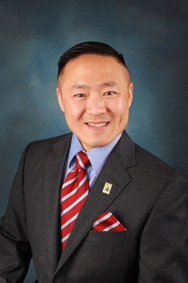 City Attorney Steven Wang wearing a dark gray suit with a blue collared shirt and a red and blue striped tie and handkerchief with a city of folsom pin for a Formal Portrait Headshot with a dark blue background