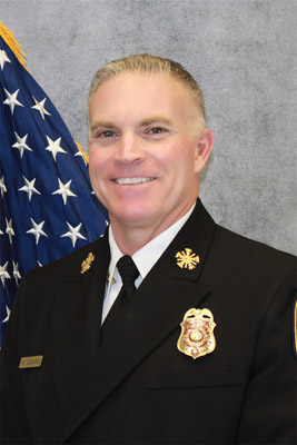 Fire Chief Ken Cusano professional portrait headshot with a light gray cement background and an American flag, wearing a black jacket, a white button-up, and a black tie with a gold name tag, gold pins, and a badge