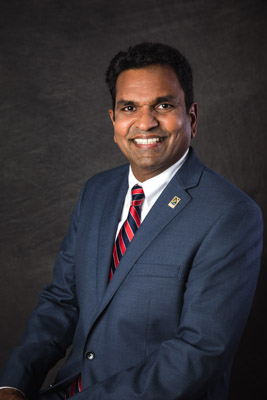 Councilmember YK Chalamcherla professional portrait headshot on a dark gray background, wearing a navy jacket with a white button up and a red and blue striped tie and City of Folsom pin