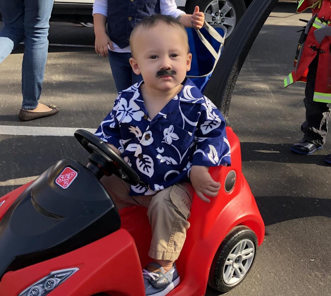 Toddler boy dressed in Hawaiian shirt with fake mustache rides in toy car