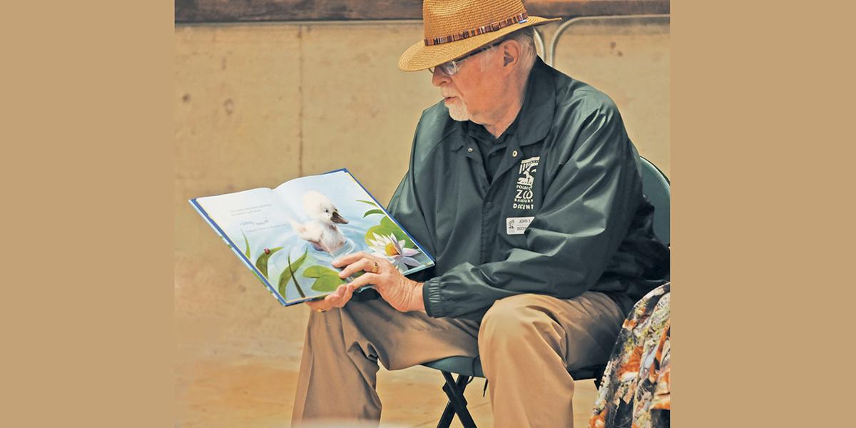 Folsom Zoo Sanctuary Docent wearing a jacket with brown slacks and a brown woven hat reading a children's book on a page with a white duck in the water