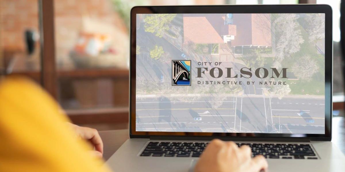 someone wearing a yellow shirt looking at the laptop screen of city of folsom side by side colored logo with a white overlay on top of an aerial shot of city hall