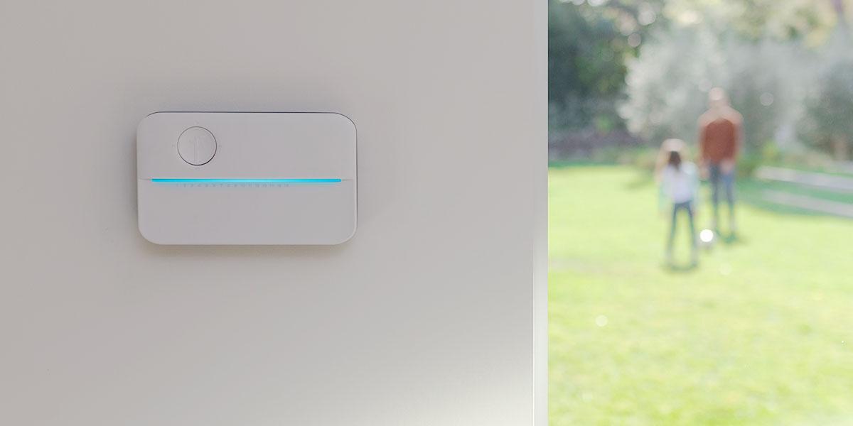all white water wifi device with a blue light handing on a white wall with a dad and child in the background playing on the grass