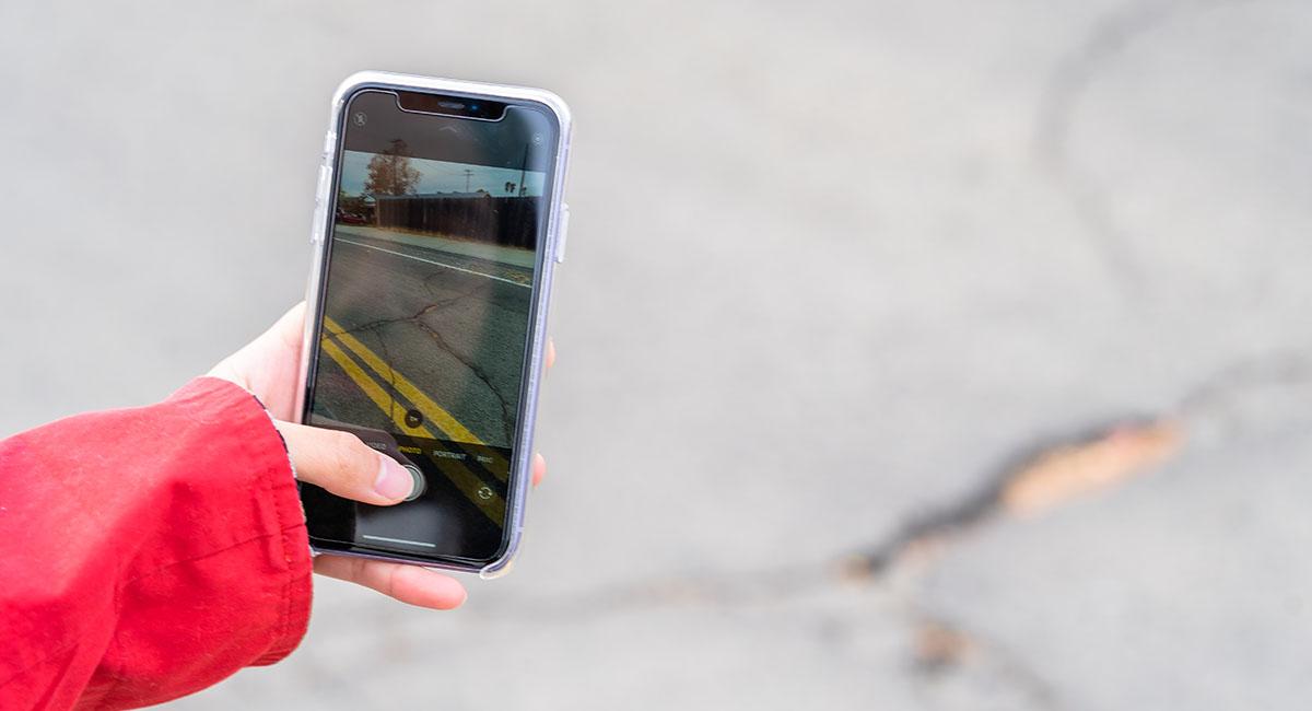 see click fix photo of someone wearing a red jacket taking a photo of a crack/ditch/pothole in the street to post onto SeeClickFix