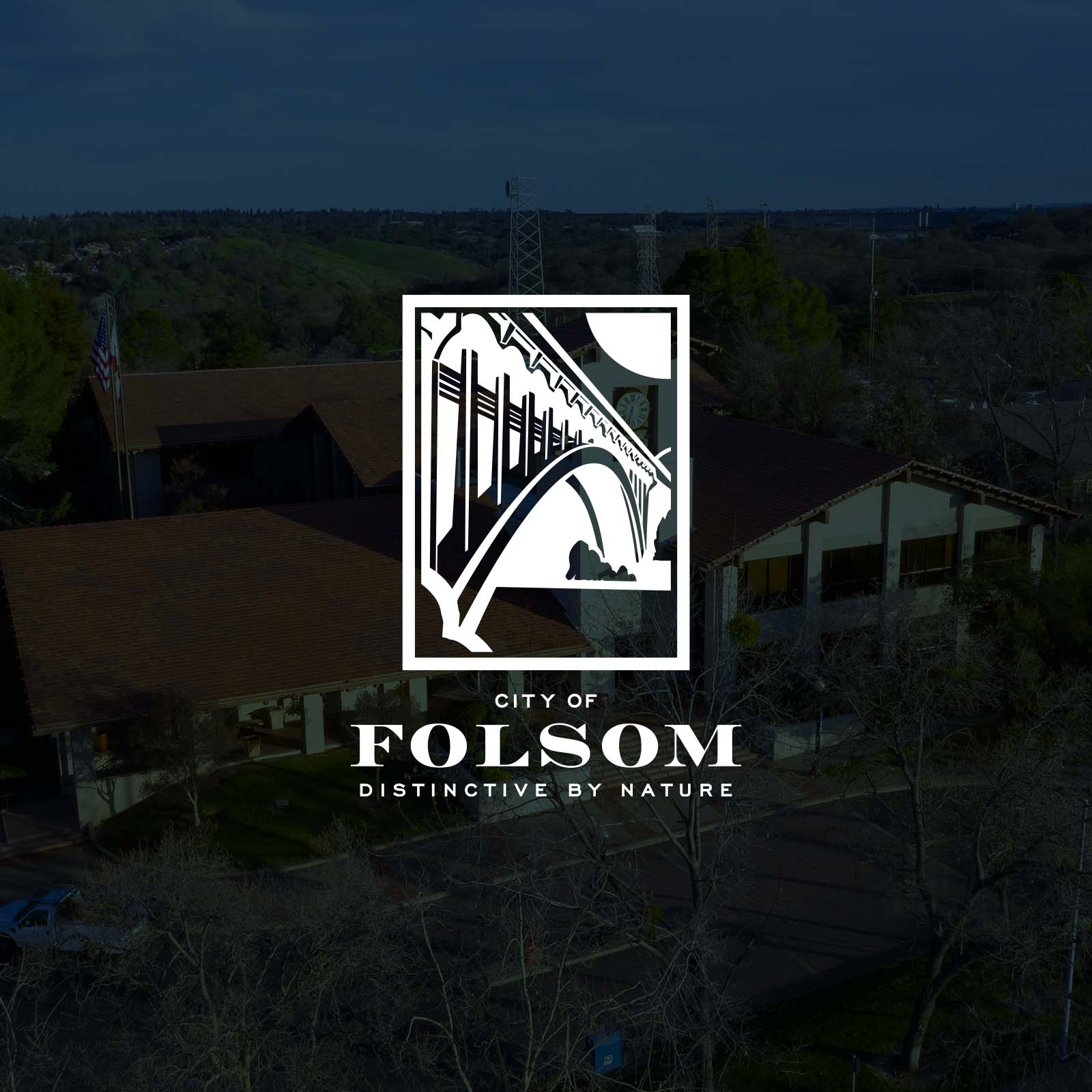 Folsom Social Graphic with white Logo with a dark overlay on top of a drone overhead shot of city hall with the grassy hills in the background
