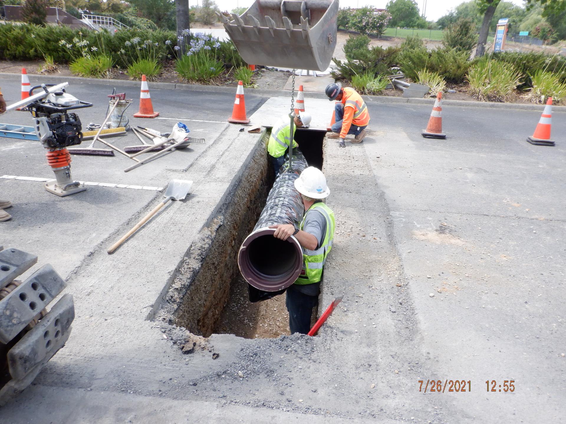 Street workers fixing an underground pipe on a road with construction tools, cones, and three workers with hard hats and reflective vests. Plants and grassy areas in the background 