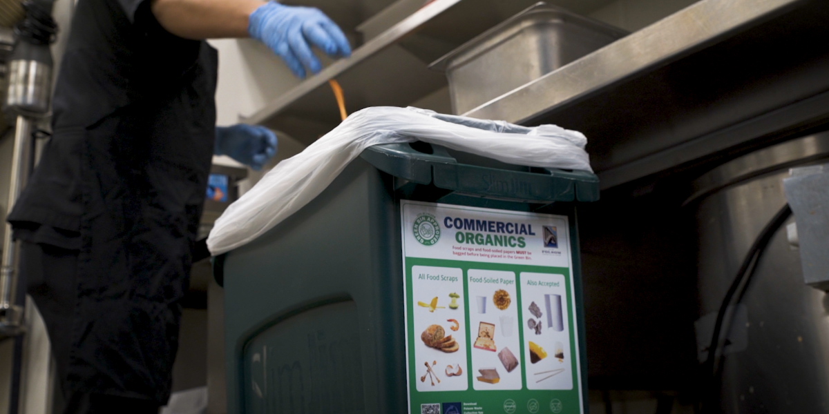 organic waste bin with a food service worker in the kitchen wearing all black with blue food latex gloves throwing waste into the commercial organics bin