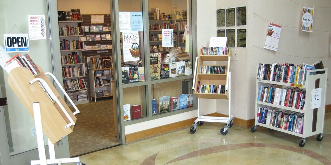 bookstore front at library with an open sign on the door and 3 shelves filled with various books for sale