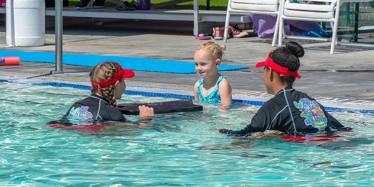 SeaSquad swim lesson of two instructors with a female student wearing a blue swimsuit, with instructors wearing the sea squad rash guard and red visors showing the student how to use the paddle board