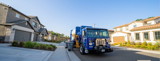 Blue Solid Waste vehicle picking up organic recycling in a residential neighborhood on a sunny day with a clear blue sky