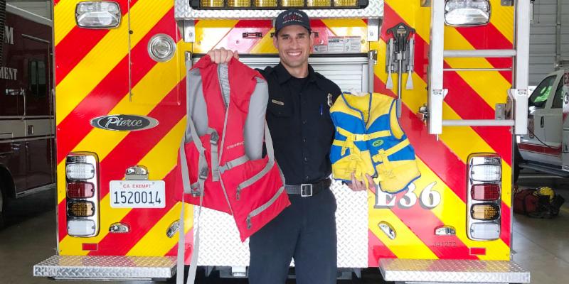 Life jacket fire department program picture of a firefighter holding an adult size red and gray life jacket and a blue and yellow kid size of life jackets in front of the back of a yellow and red striped fire truck