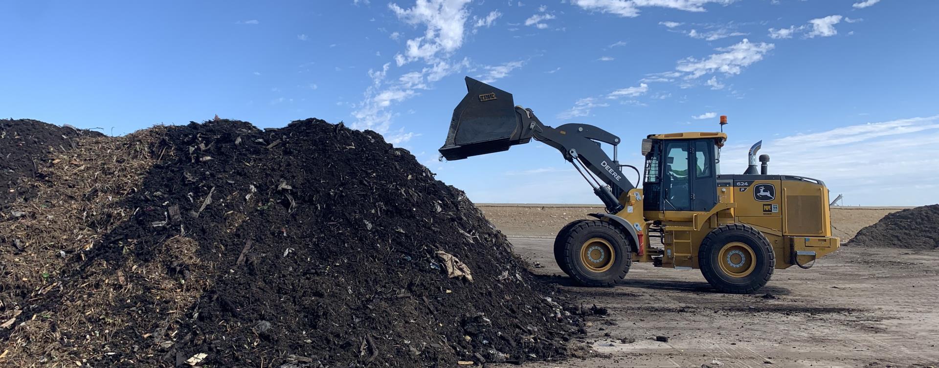 Large pile of compost outside on concrete pad with blue sky in the background. A tractor beside the pile has its bucket in air as it mixes the compost.
