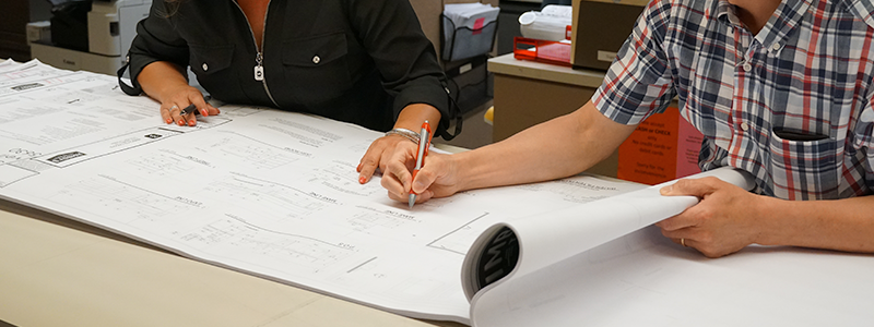 Development Engineering Services page header showing two employees upper torsos analyzing the engineering map with pens in hand and pointing to the paper.