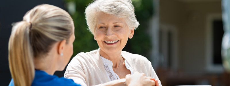 Seniors Helping Seniors page header. Stock image of a senior wearing a white blue and beige cardigan looking at another person wearing a blue shirt.