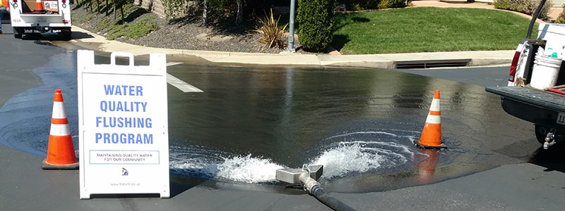 water in street with City equipment, vehicles, cones and sign for Water Quality Flushing Program